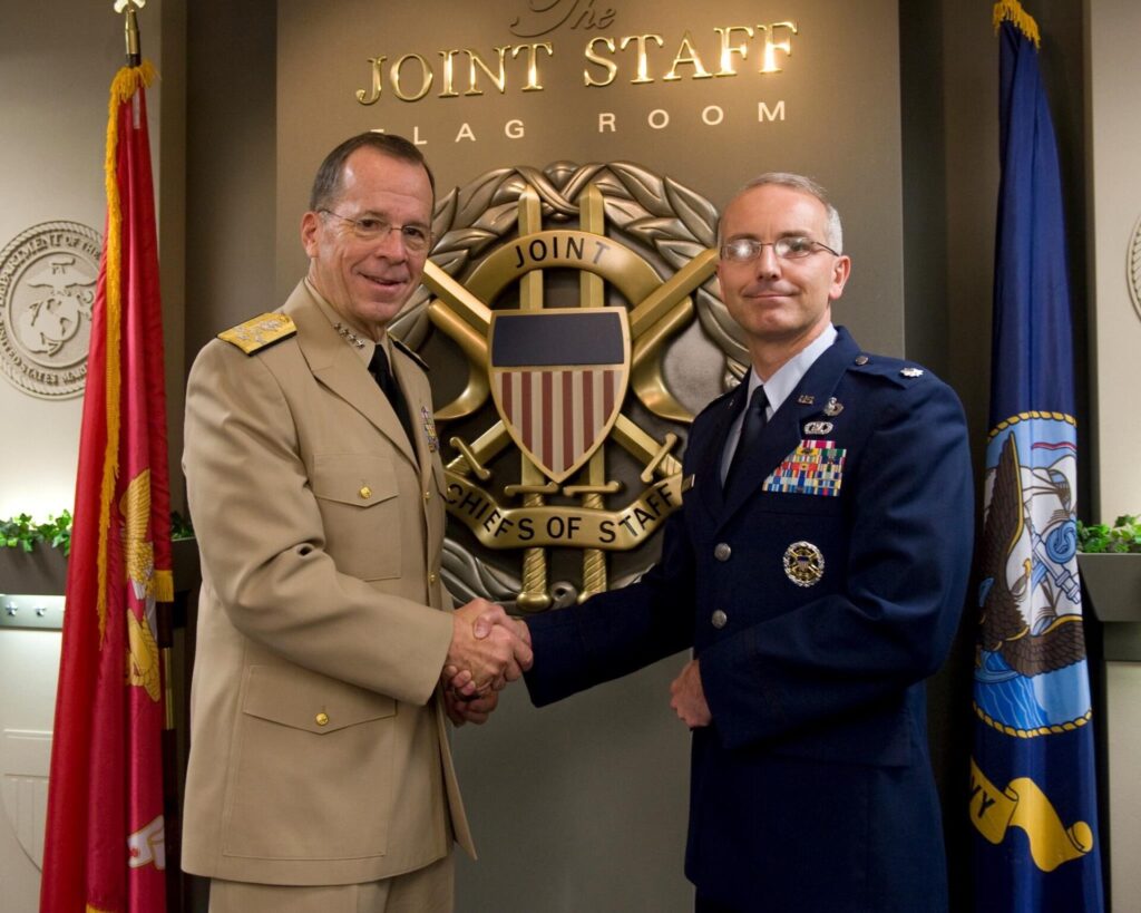 Two men in military uniforms shaking hands in front of a seal on the wall that reads "The Joint Staff Flag Room".